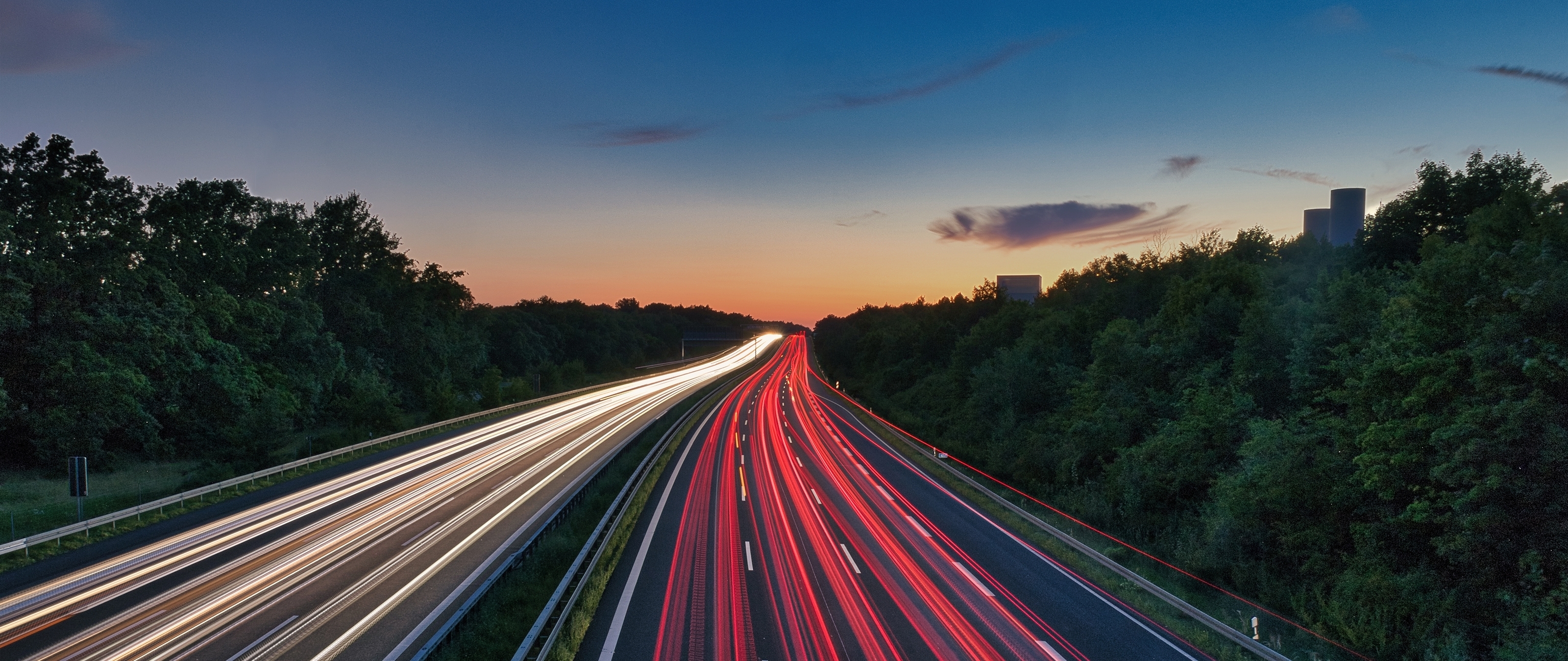 Long-exposure photo of a highway at dusk with light trails from vehicles, surrounded by trees and a vibrant sky.